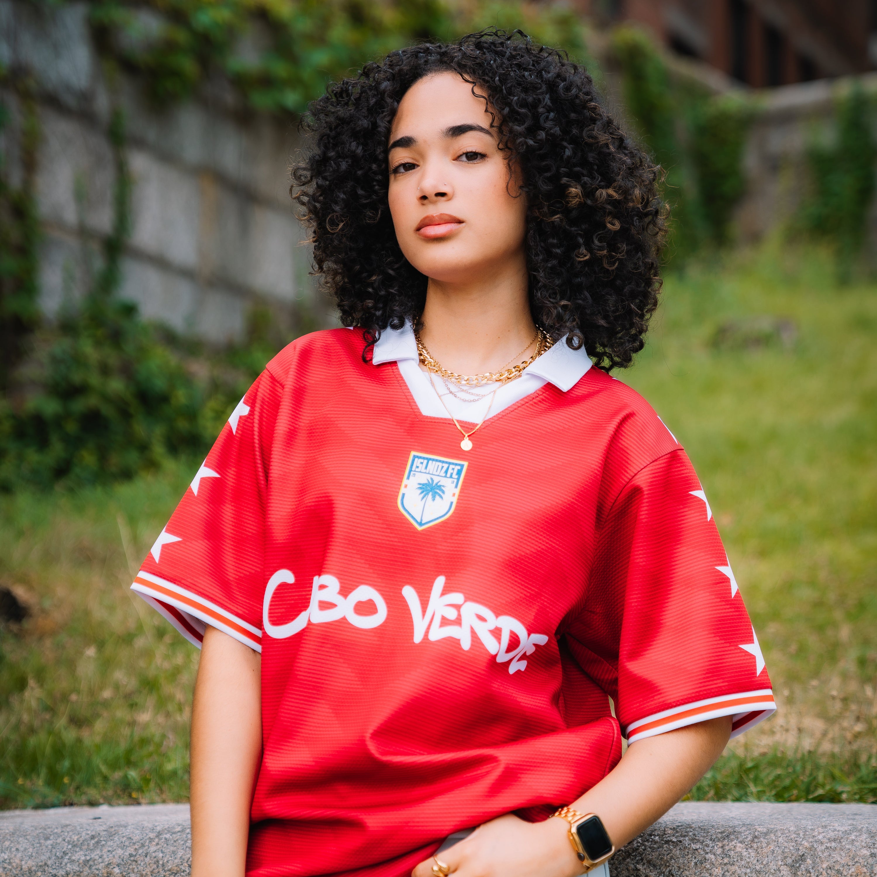 A woman with curly black hair stands outdoors wearing a red short-sleeve soccer jersey with white lettering and a white collar, paired with light blue jeans. The jersey features a blue crest on the chest and 'CBO VERDAD' printed across the front.