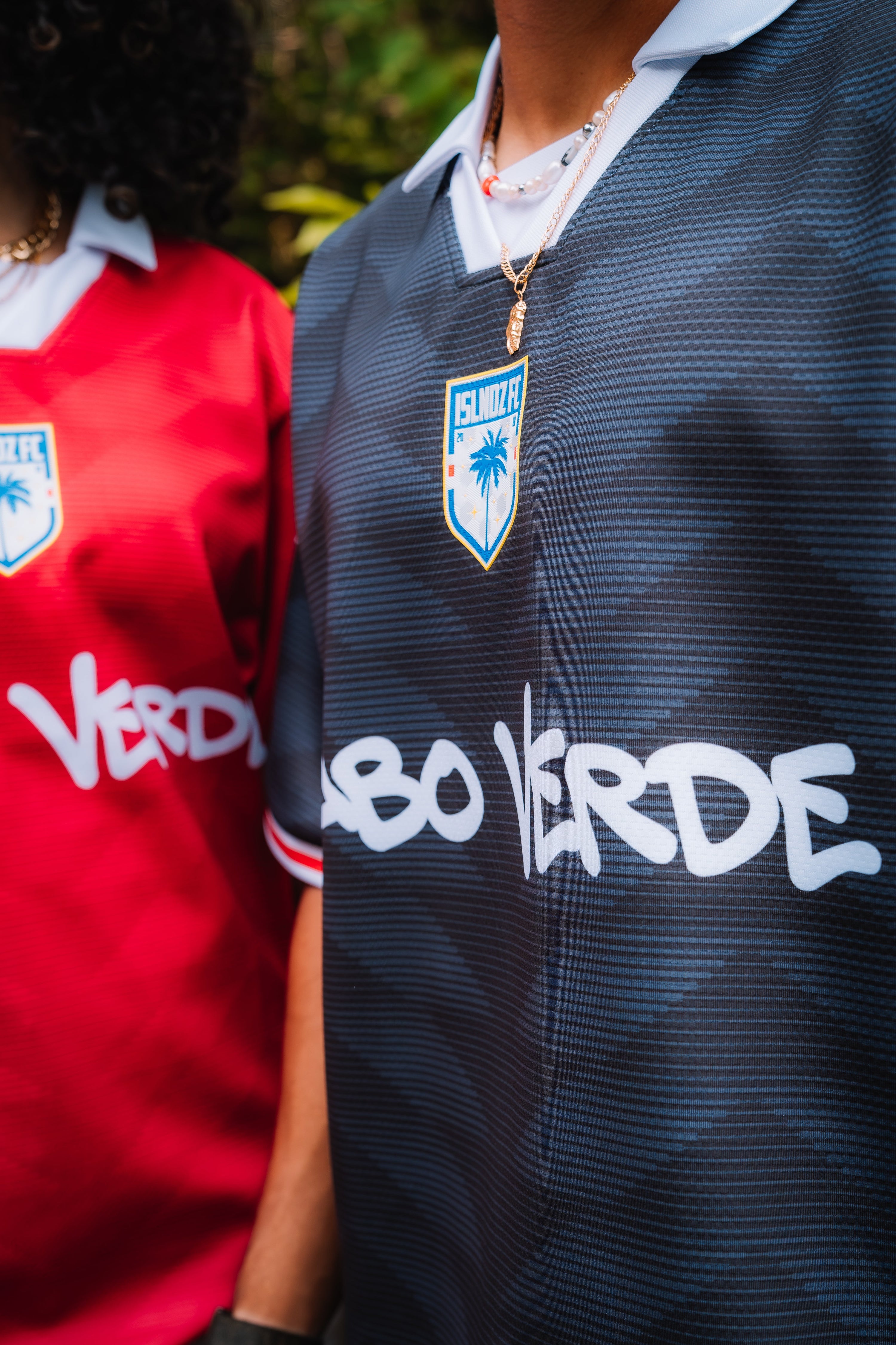 Close-up of a person wearing a dark navy textured soccer jersey with a blue and white shield crest and large white 'CABO VERDE' text across the chest, standing outdoors next to another person in a red jersey.