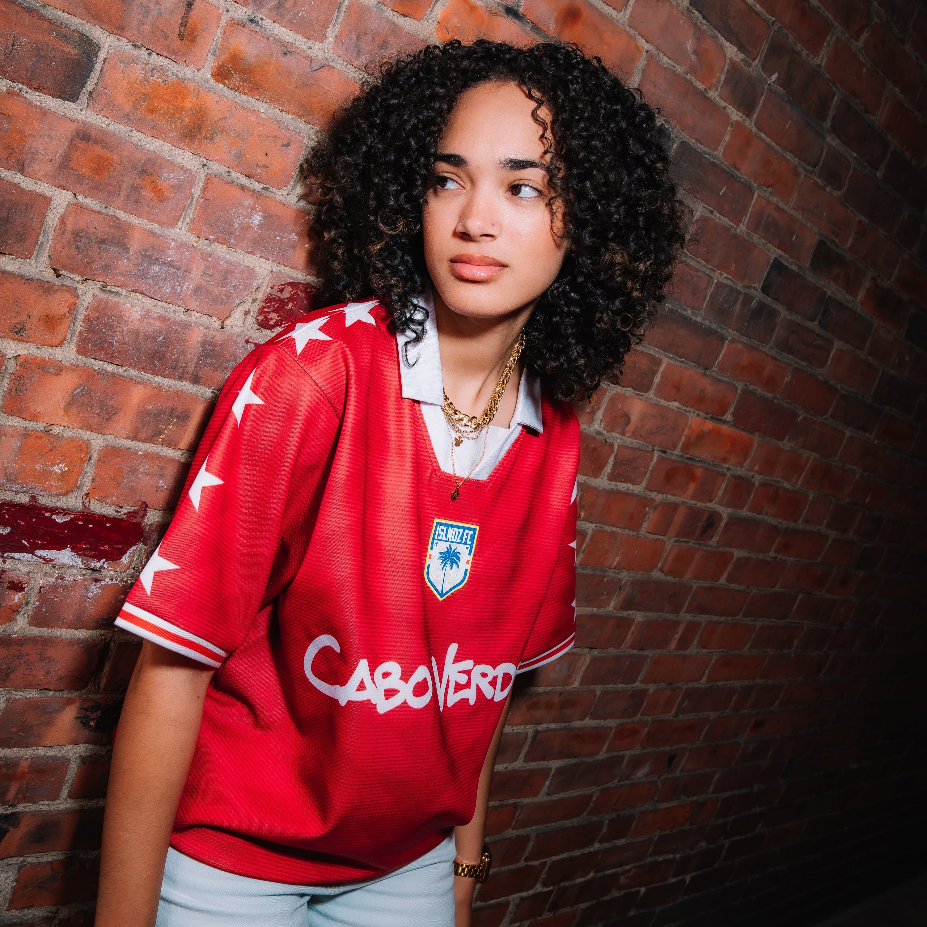 A woman with curly black hair stands against a red brick wall wearing a red short-sleeve soccer jersey with white trim, a white collar, a blue crest, and the name 'CABOVERO' printed across the front, paired with light blue jeans.