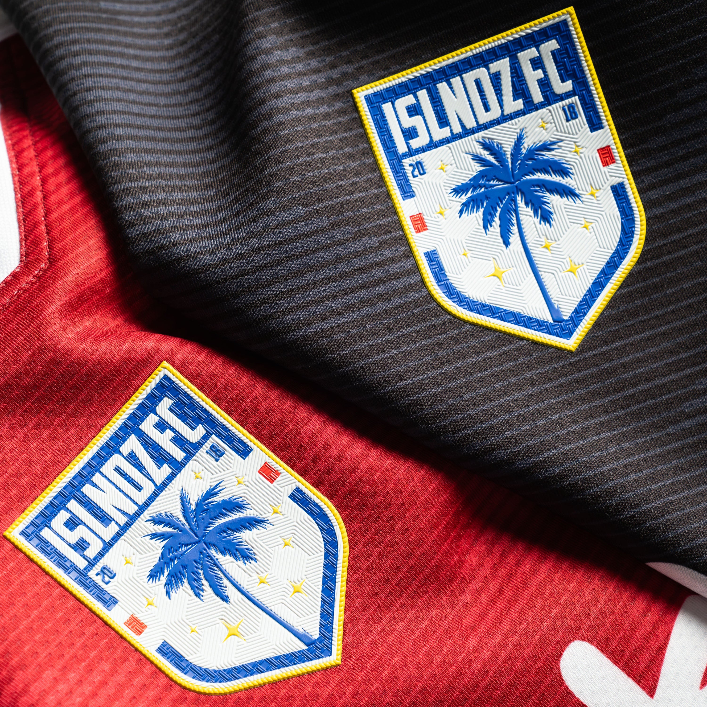 Close-up of a soccer jersey showing two embroidered ISLNOZ.FC team patches on a split red and black striped fabric background.