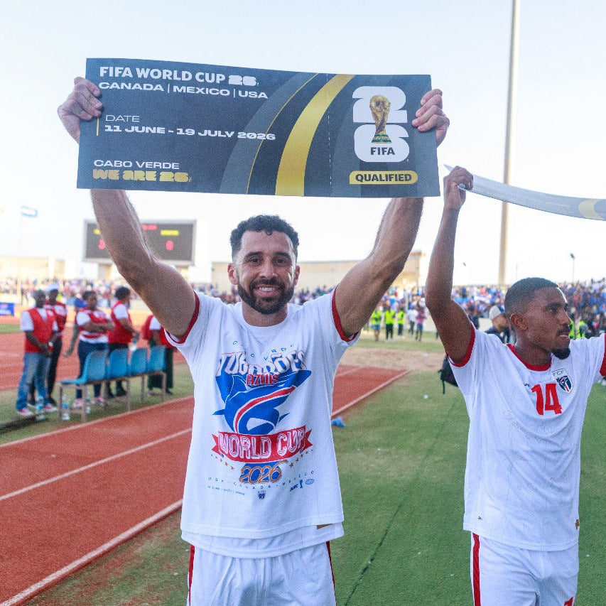 Two men in sports uniforms holding a FIFA World Cup qualification sign on a sports field.